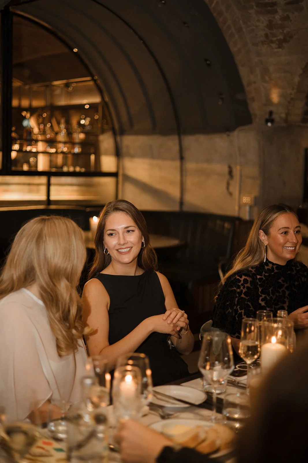 Women socializing at a dinner event in a dimly lit restaurant with candles, multiple wine glasses, and elegant attire.