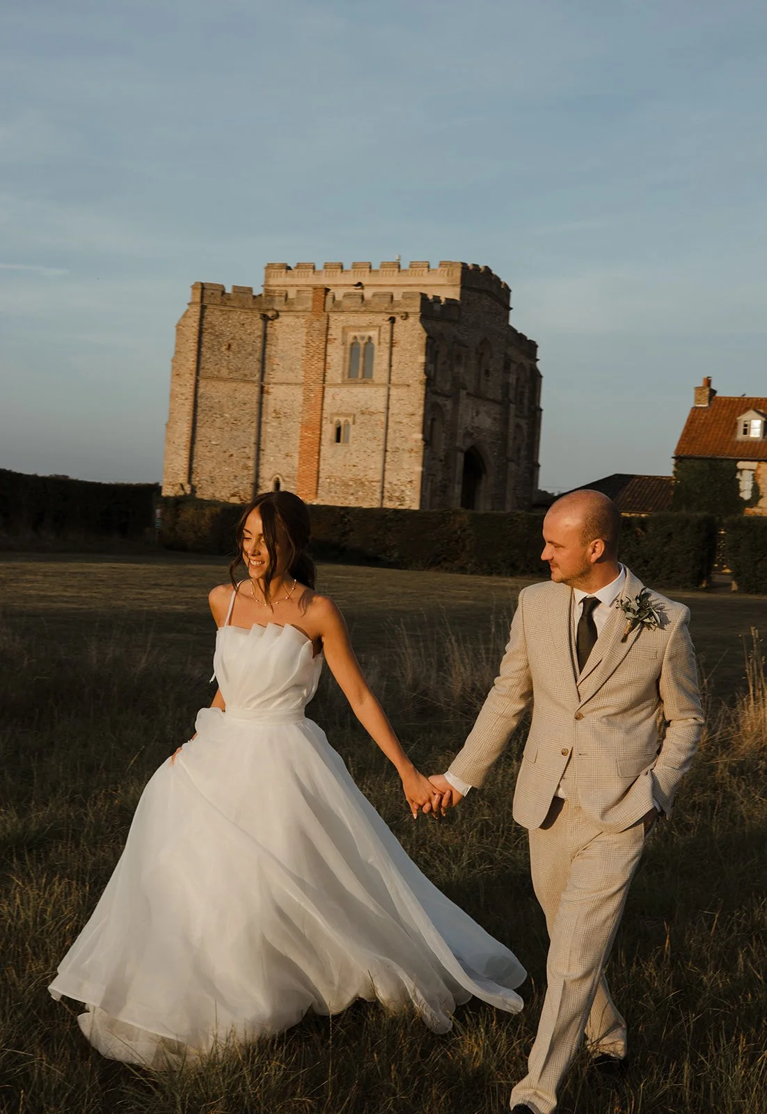 A bride and groom holding hands and walking in a grassy field at sunset, with a historic stone castle in the background.