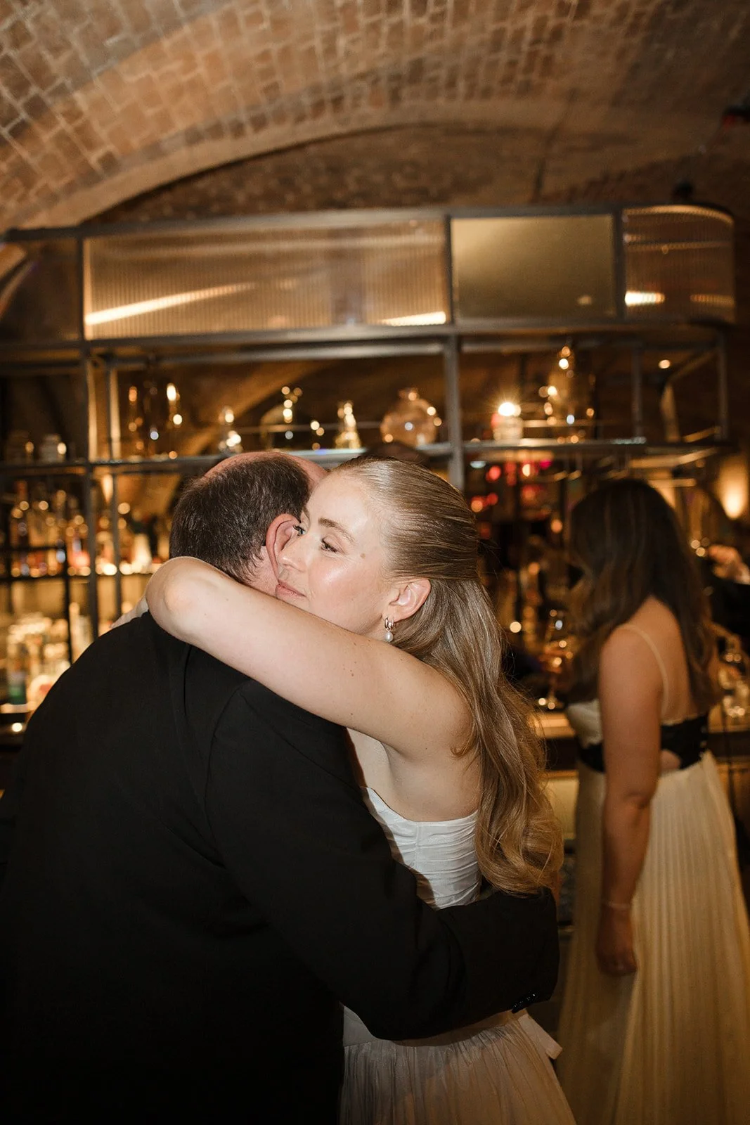 A woman and a man hugging warmly in a dimly lit bar or restaurant with a brick vaulted ceiling. The woman has long, wavy blonde hair and is wearing a white dress, while the man is dressed in a black suit. There are other people and bar shelves with b