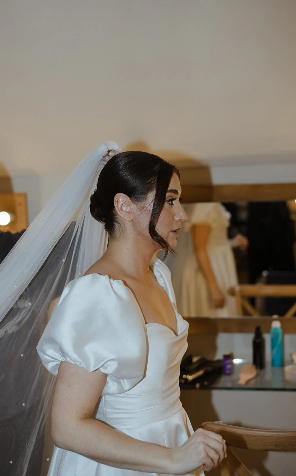 A bride in a white wedding gown with puffed sleeves and a v-neckline, wearing a veil, is standing sideways in a room, possibly a dressing area, with beauty and hair products visible on a table behind her.