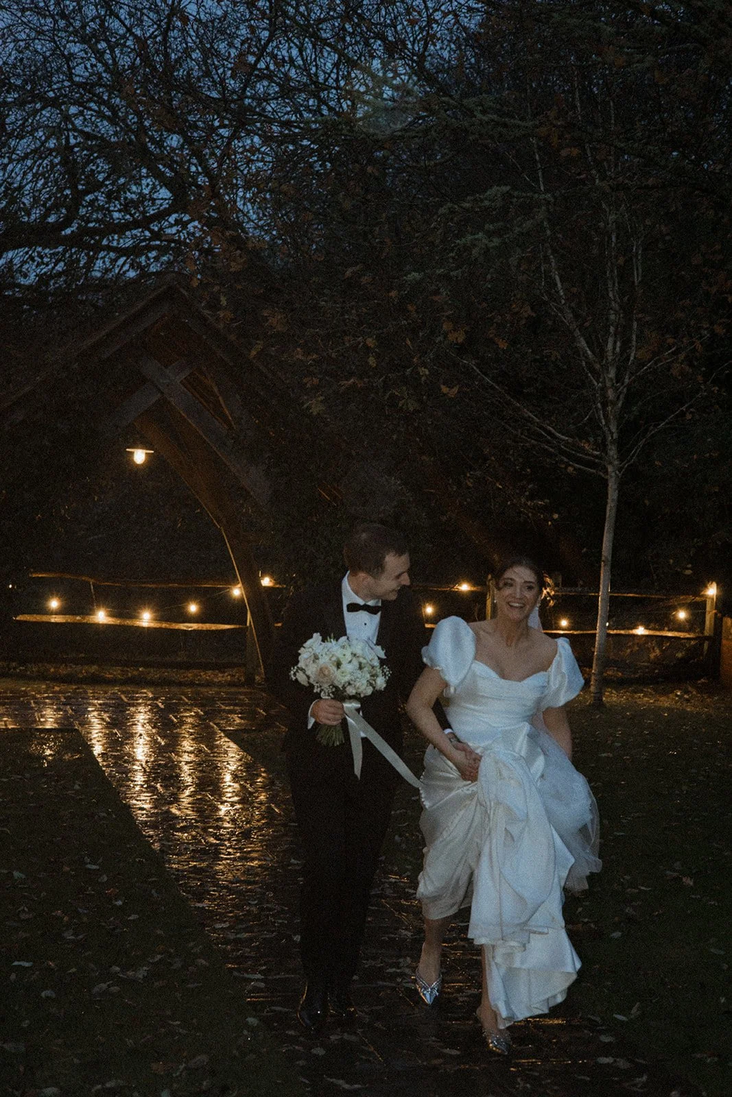A bride and groom walking together outdoors at night, the groom in a tuxedo holding a bouquet, the bride in a white gown lifting her dress slightly, under string lights with trees and a wooden structure in the background.