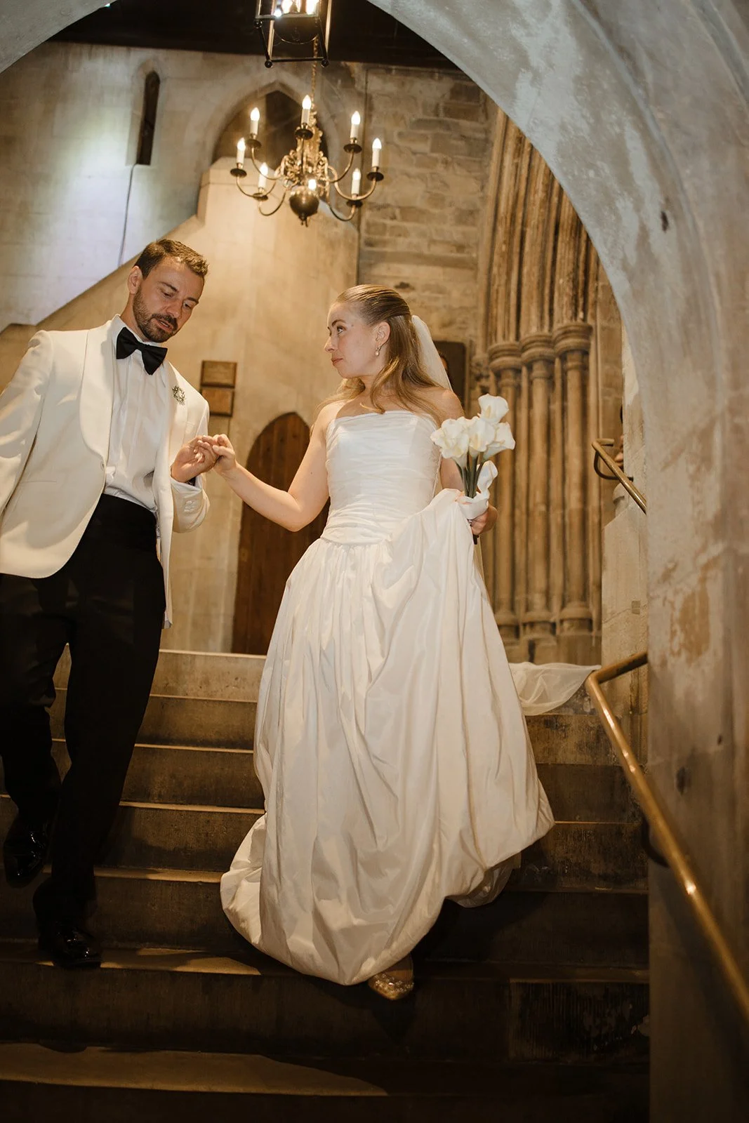 A bride and groom descending a stone staircase in a church, holding hands, with the bride holding a bouquet of white flowers. The bride is wearing a strapless white wedding gown, and the groom is dressed in a white tuxedo jacket with black pants and 