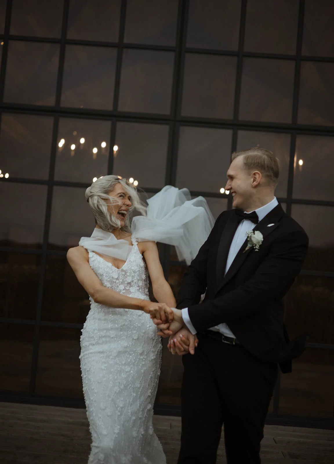 A bride and groom holding hands and smiling at each other indoors, with large windows and chandeliers in the background.