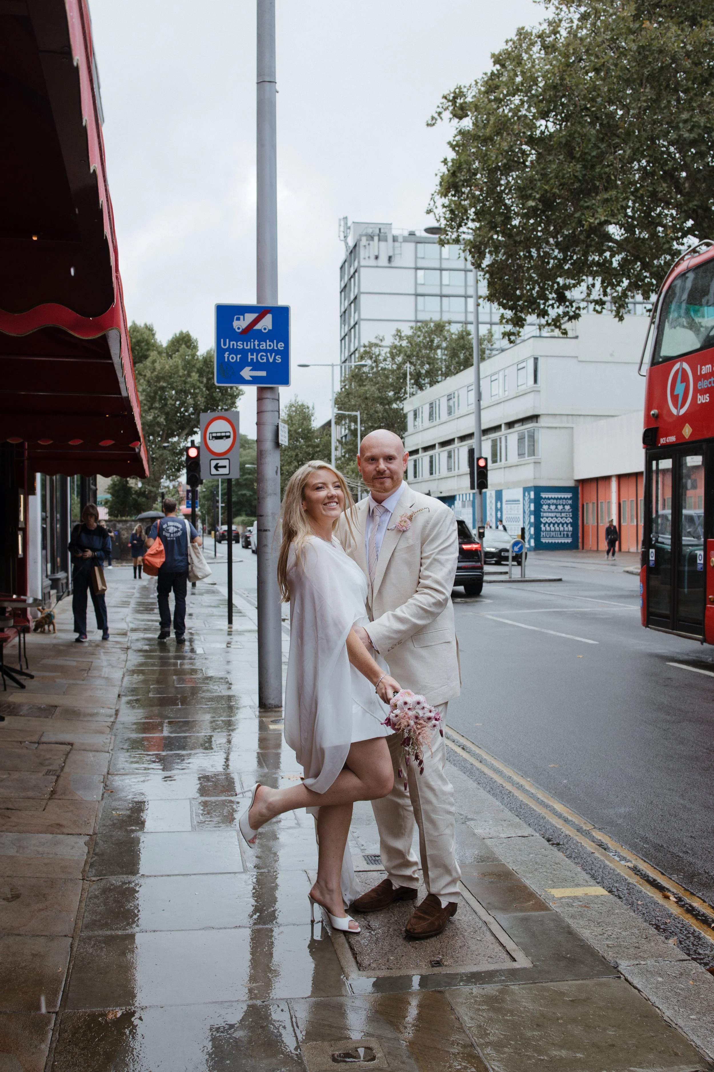 A newlywed couple standing on a rainy city sidewalk, smiling happily; the bride is kneeling on one leg, holding a bouquet, wearing a white dress and high heels, while the groom is standing beside her, dressed in a beige suit and brown shoes.