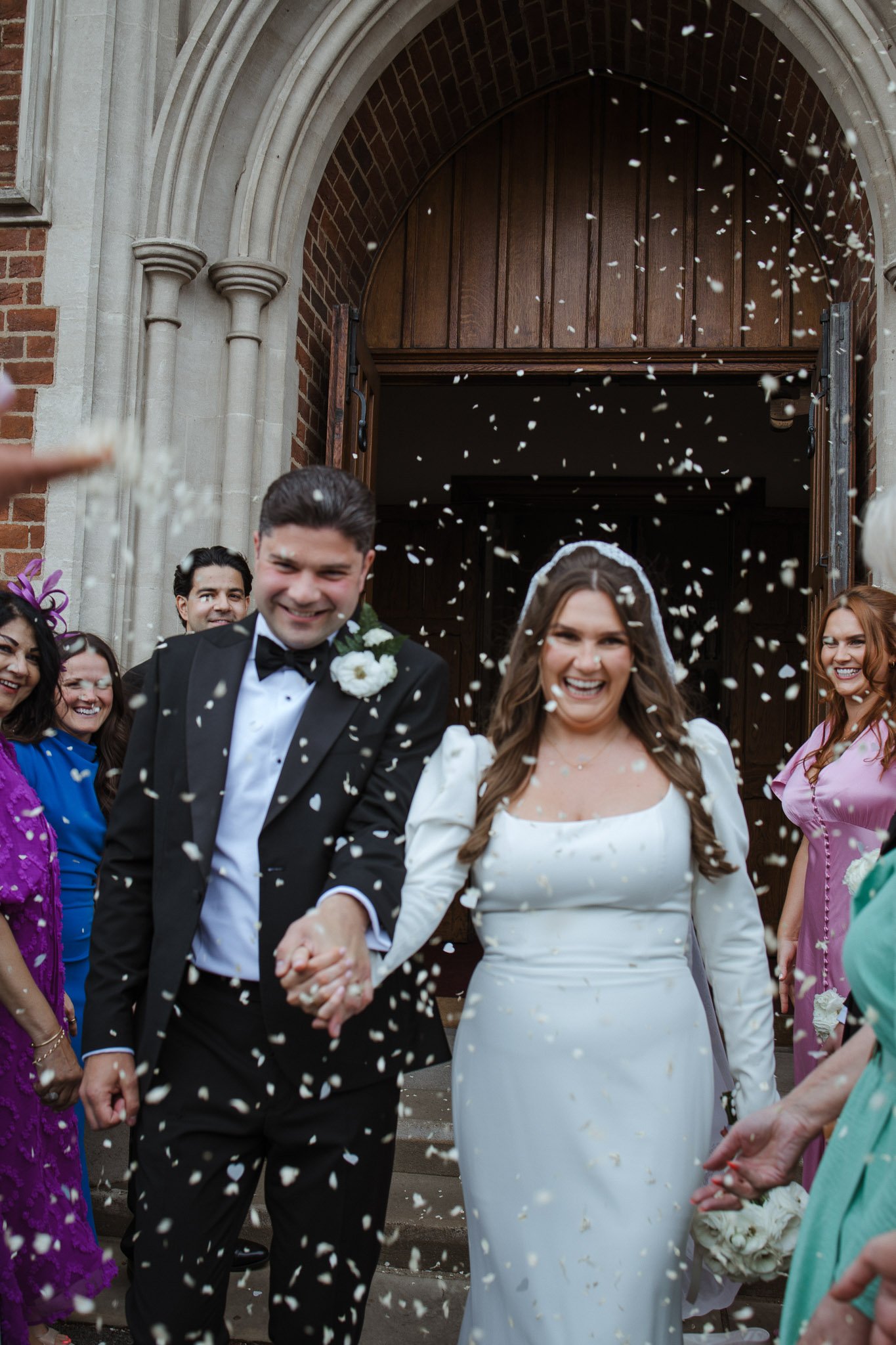 A newlywed couple walking out of a church hand in hand, smiling, as guests throw confetti around them.