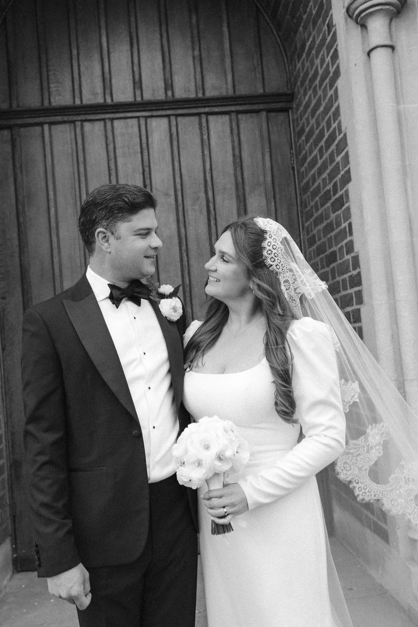 Black and white photo of a bride and groom standing close, looking at each other and smiling. The bride is wearing a white dress with a veil and holding a bouquet of flowers. The groom is dressed in a tuxedo with a bow tie. They are standing in front