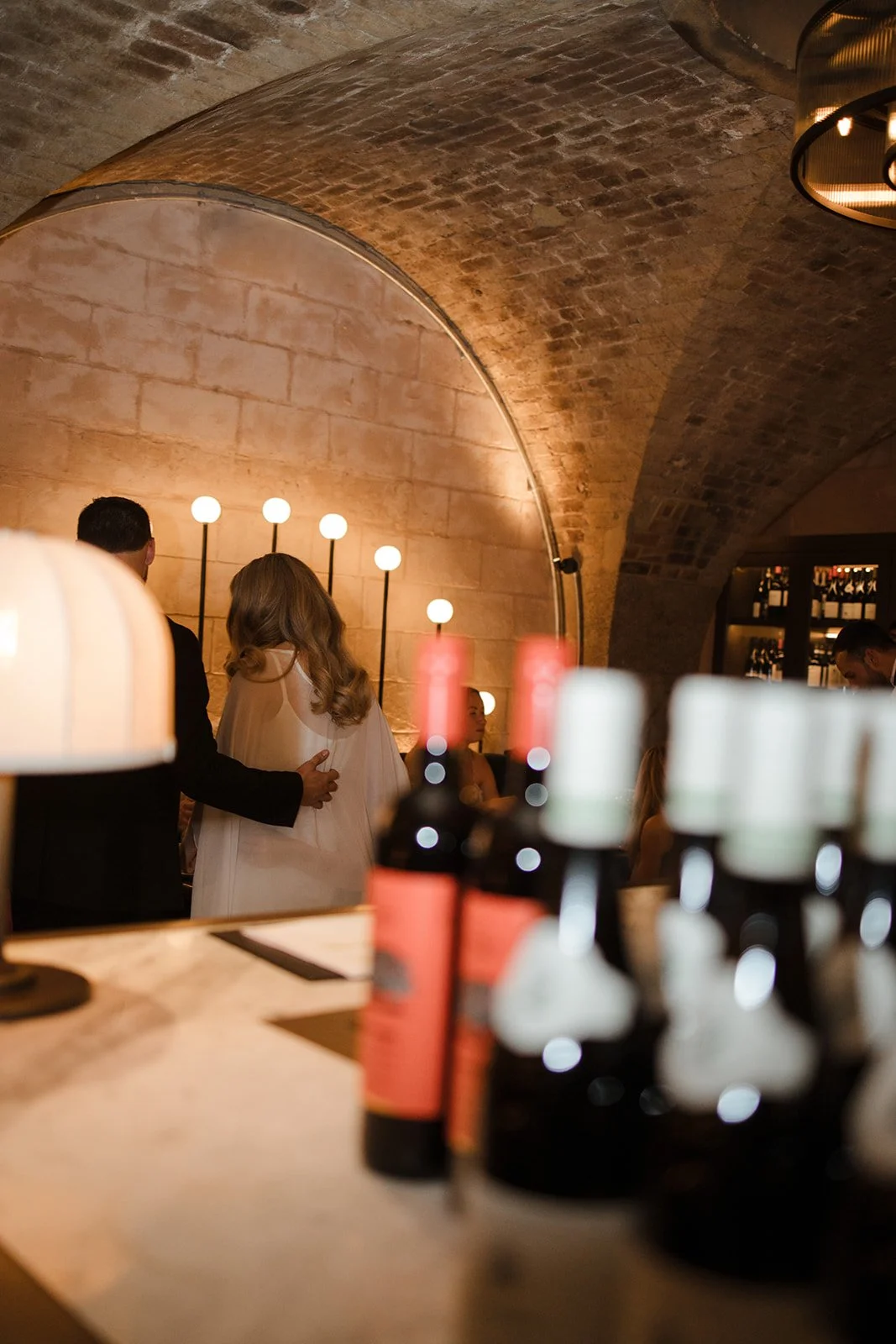 A cozy bar or restaurant with a brick arched ceiling, warm lighting, and bottles of wine on the shelves. People are gathered, possibly celebrating, with some couples standing close together.