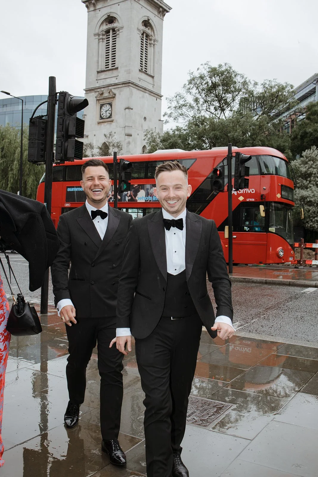 Two men in black tuxedos and bow ties walking on a rainy city street, smiling, with a red double-decker bus and a historic clock tower in the background.