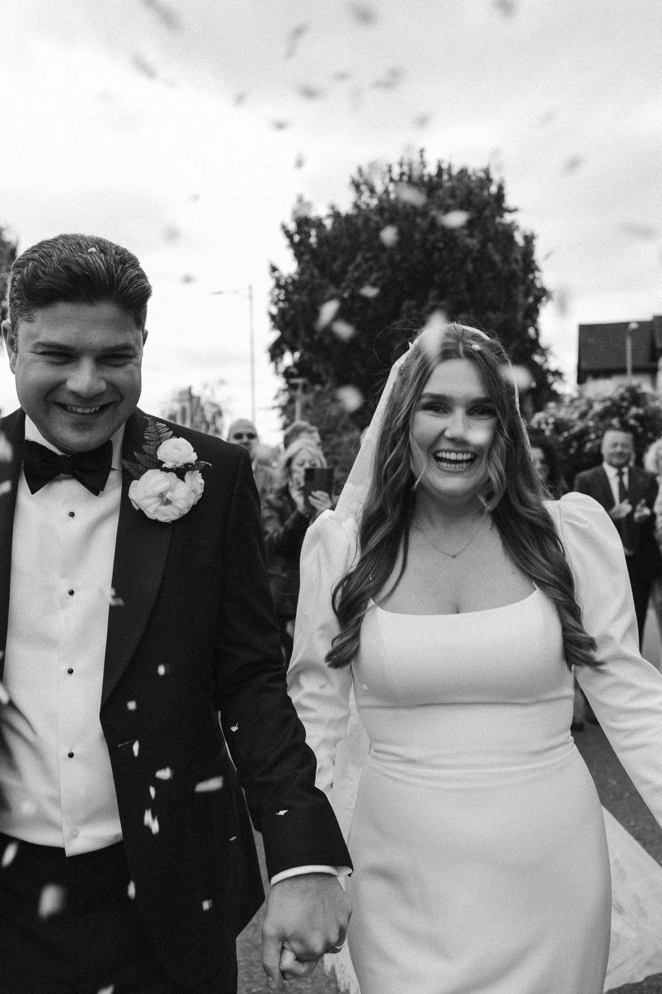 Black and white photo of a smiling bride and groom holding hands, walking outdoors at their wedding with guests in the background.