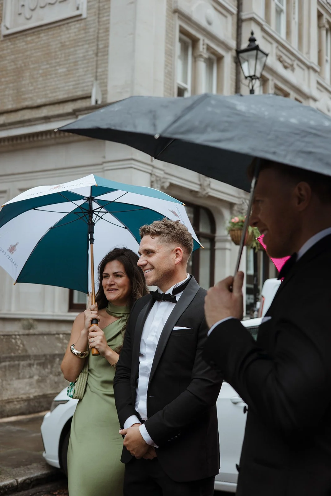 A group of three people, two women and one man, standing outside on a rainy day, holding umbrellas. The man is dressed in a tuxedo, and the woman in the green dress is smiling. They appear to be at a formal event.