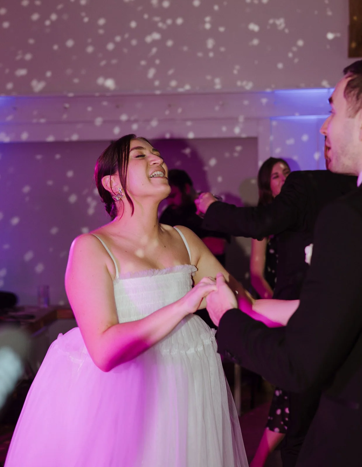 A woman in a white dress dancing and smiling with a man in a black suit at a party or celebration, with others in the background and disco lights creating a festive atmosphere.