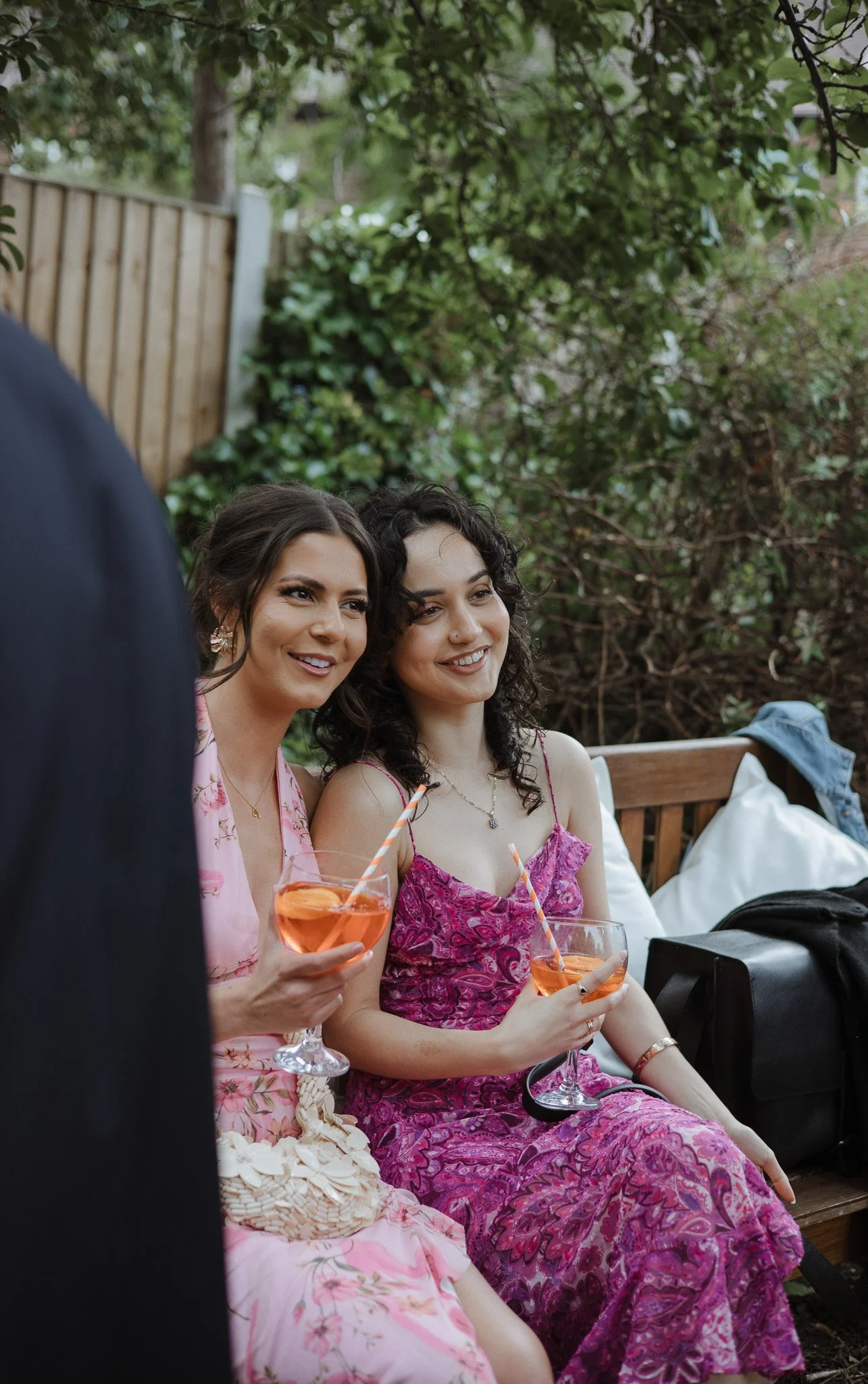 Two women sitting on a wooden bench outdoors, smiling and holding pink cocktails with orange slices and striped straws, with greenery and a wooden fence in the background.