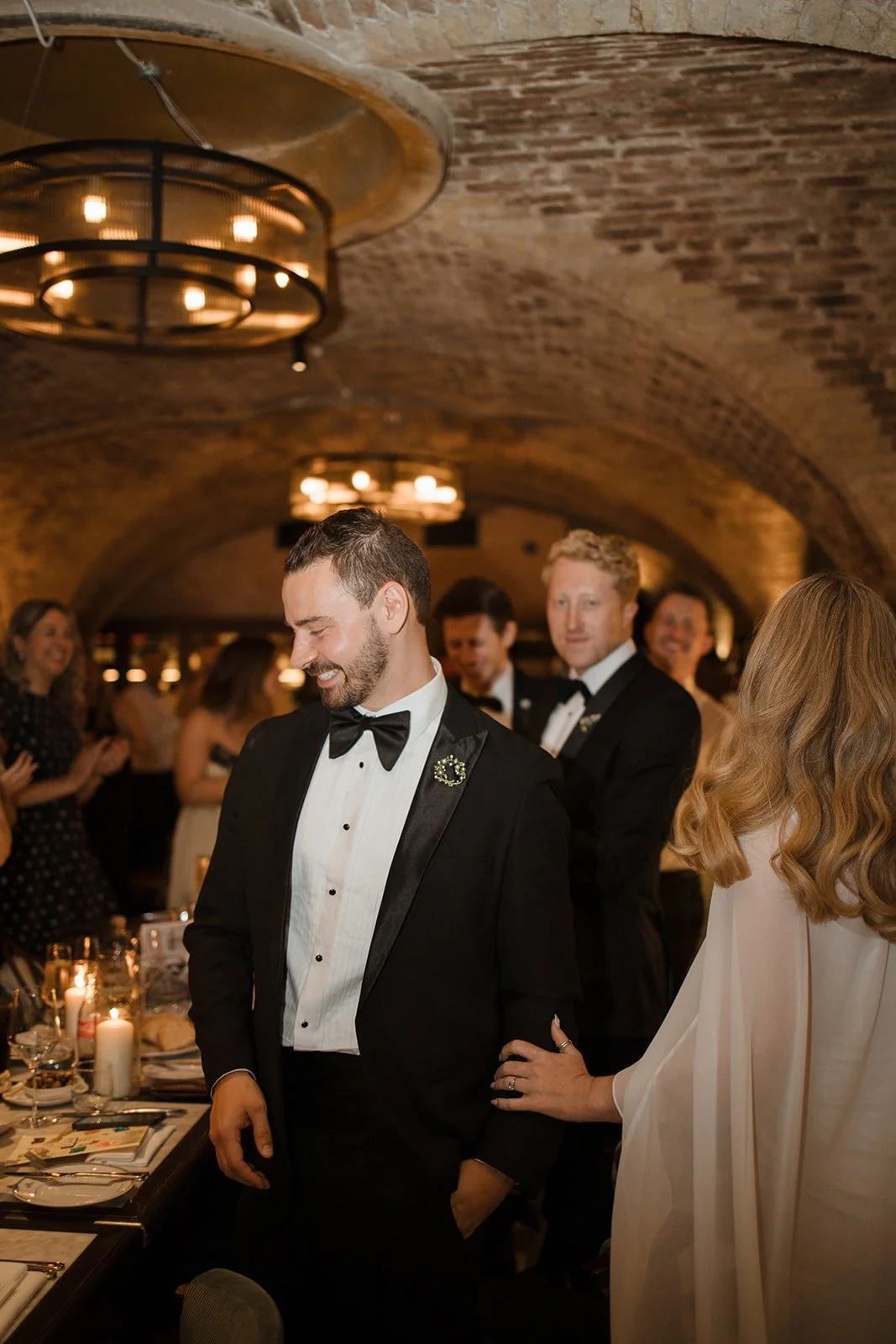 A man in a black tuxedo with a bow tie is smiling during a formal event in a brick-walled venue, surrounded by other well-dressed guests.