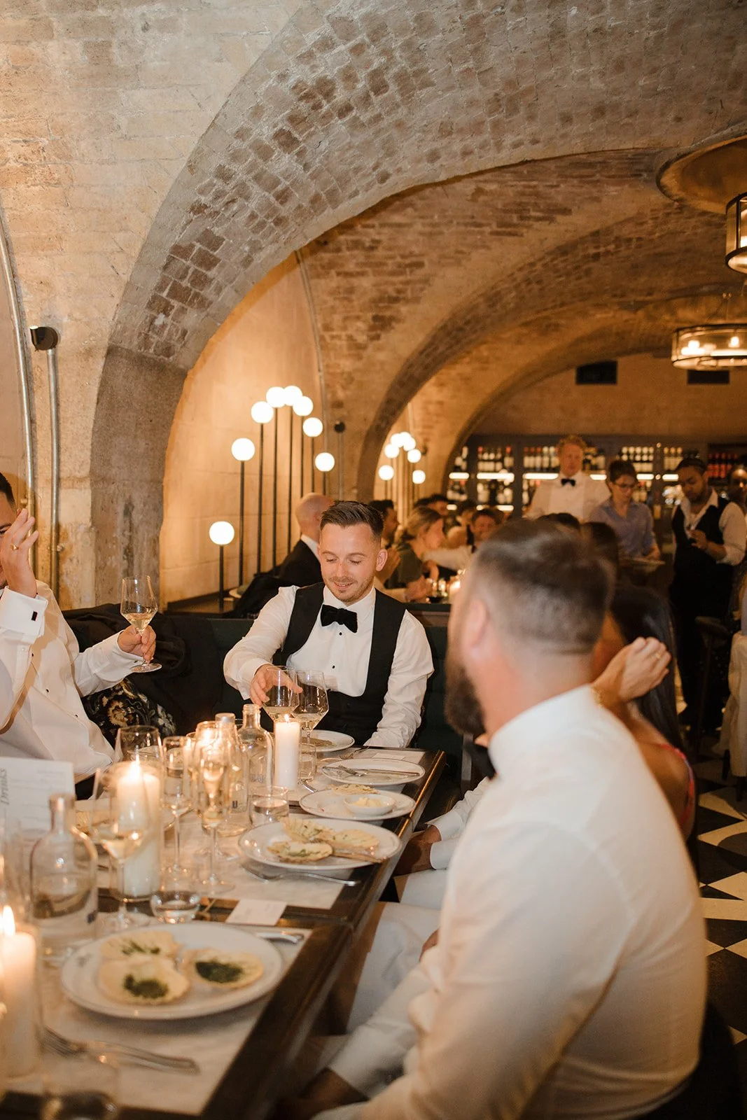 A group of people dressed in formal attire, sitting at a long dining table in a warmly lit restaurant with arched brick ceilings, dining and drinks, with some waitstaff serving in the background.