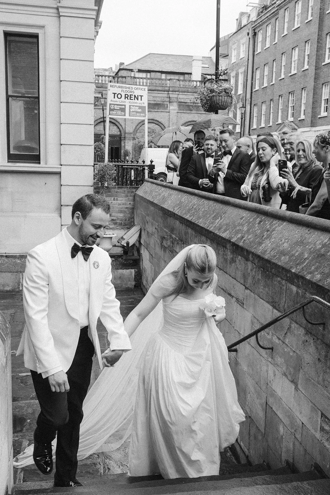 Black and white photo of a newlywed couple holding hands and walking up stairs, surrounded by a group of people taking pictures and watching outside on a rainy day.