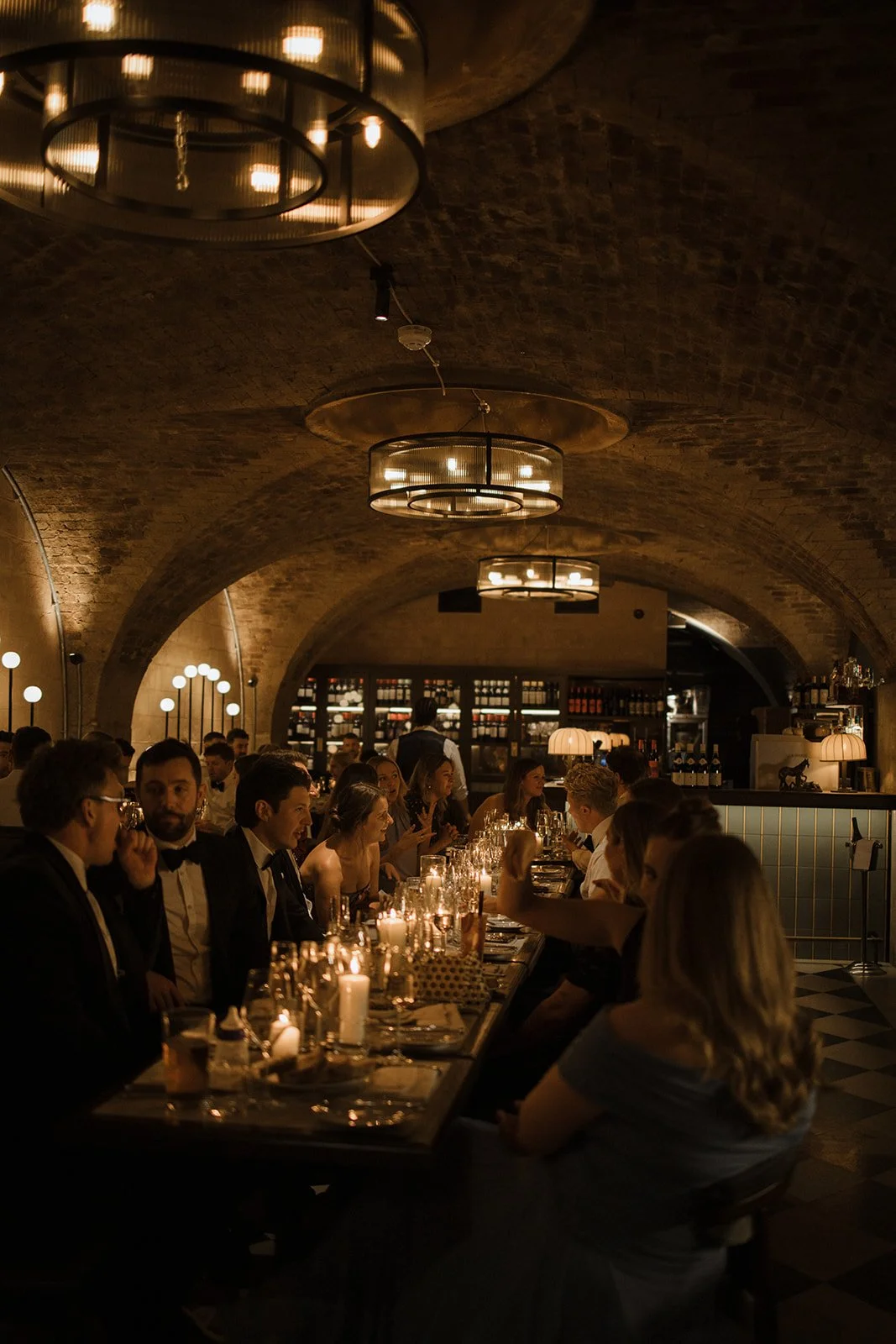 A dimly lit restaurant with a brick arched ceiling, featuring large circular chandeliers. Guests dressed in formal wear are seated at a long table, illuminated by candles, enjoying a meal. The background shows a bar with shelves of bottles and wine.