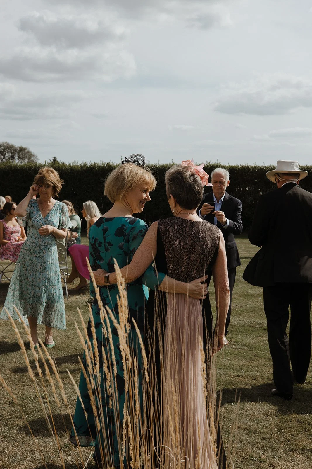 Group of people socializing at an outdoor event on a grassy field with cloudy sky background. Women are dressed in colorful dresses, and men in suits and hats.
