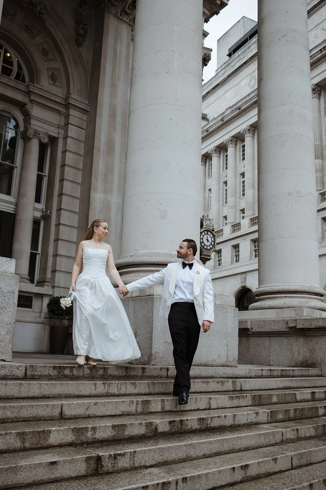 A bride and groom walking down the steps of a grand, classical-style building with large columns and ornate architecture, holding hands and looking at each other.