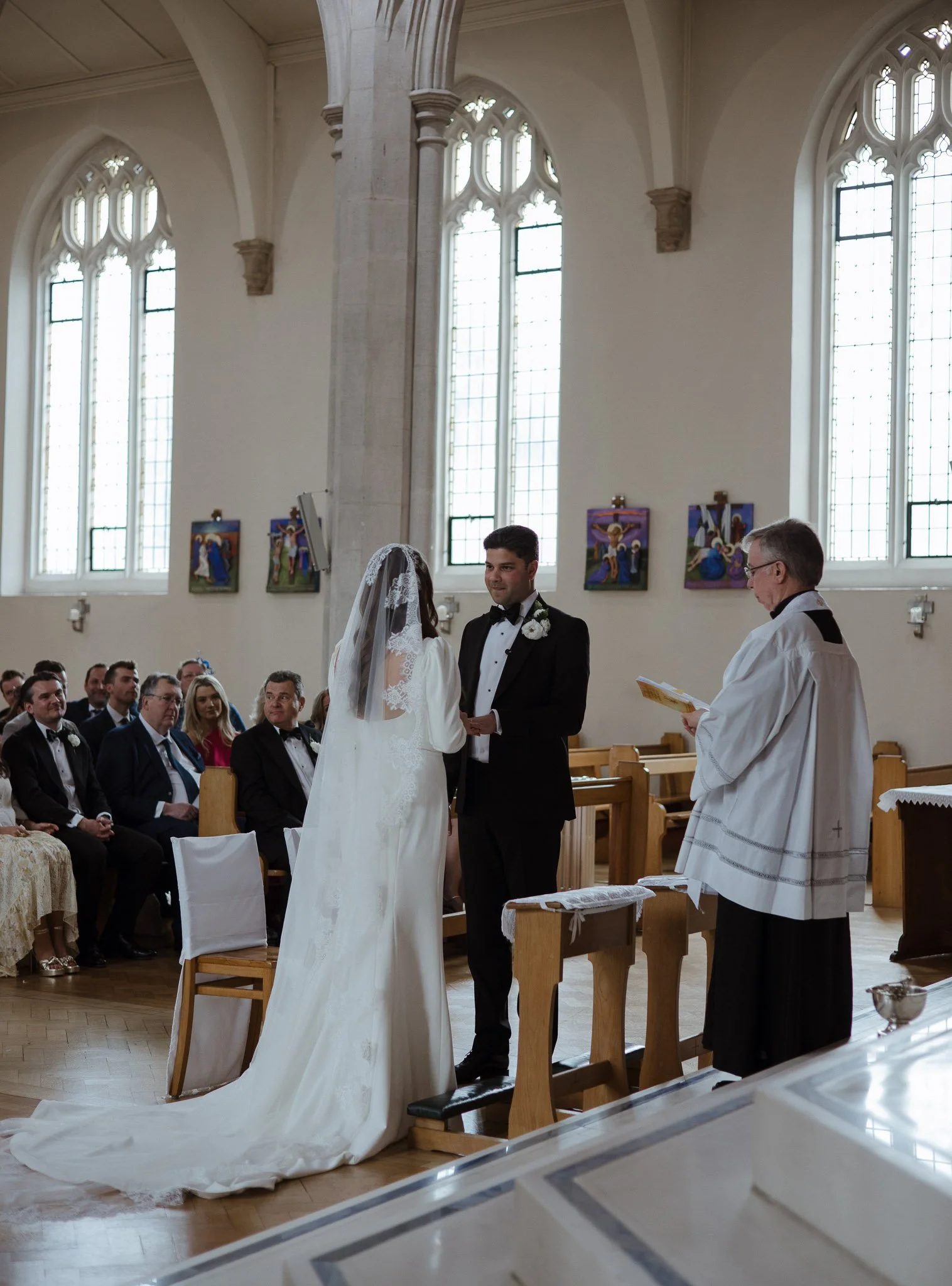 A wedding ceremony inside a church with a bride and groom standing before an officiant, exchanging vows. The bride is wearing a white wedding dress with a lace veil, and the groom is in a black tuxedo with a white boutonniere. Guests are seated in th