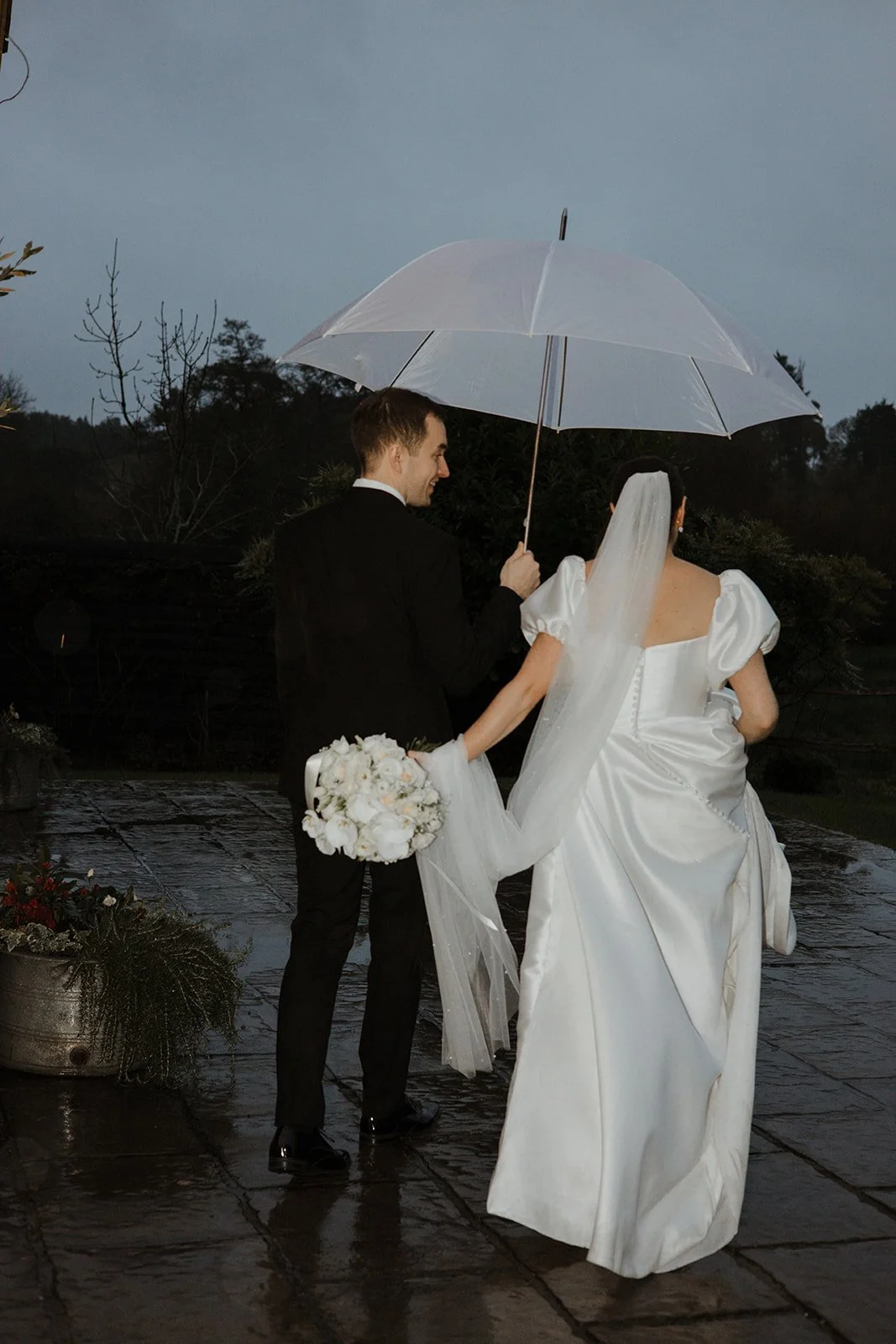 A newlywed couple walking outdoors in the rain, under a white umbrella. The groom is holding a bouquet of white flowers, and the bride is wearing a white wedding dress with puffed sleeves and a long veil. The ground is wet, and there are plants and t