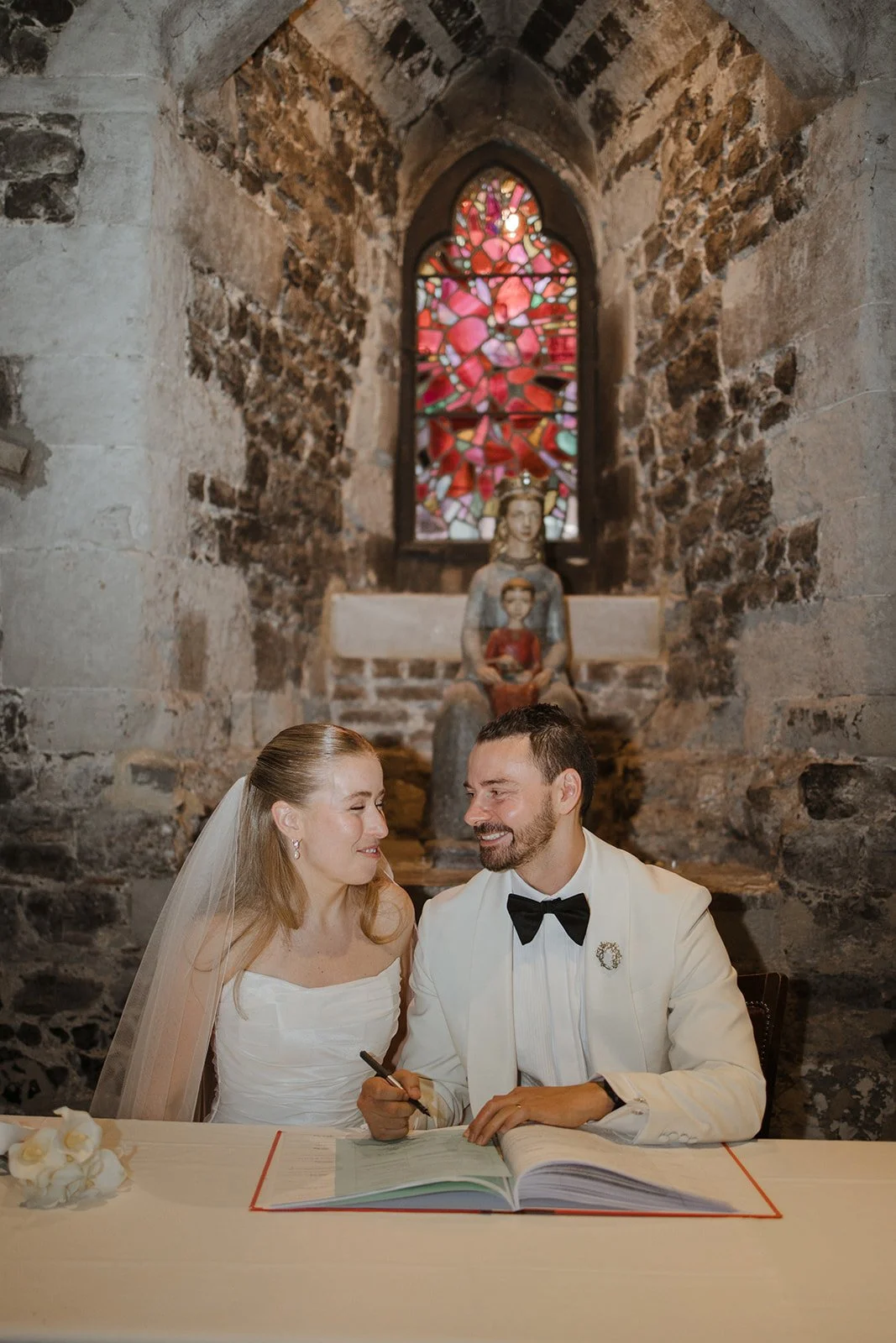 A bride and groom sitting at a table inside a stone church, signing a marriage document and smiling at each other, with a stained-glass window and religious statues in the background.