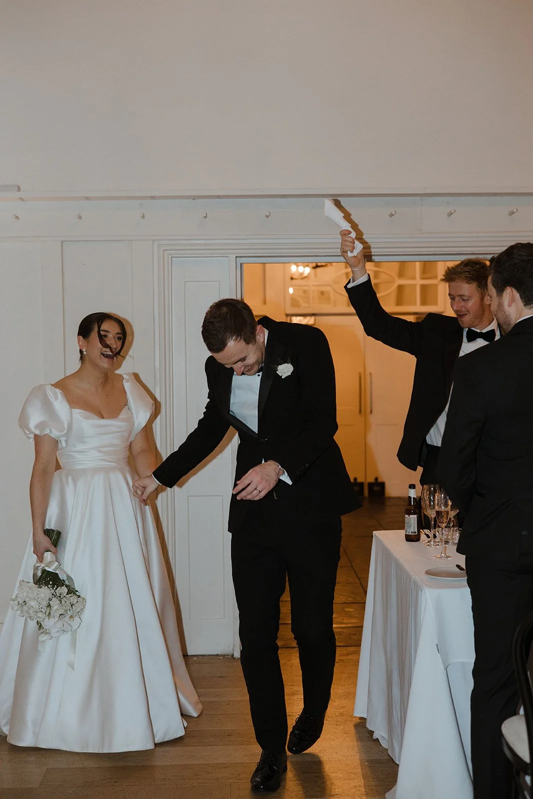 Wedding reception scene with bride in white gown holding bouquet, groom in tuxedo, and guests celebrating, one holding a napkin in the air.