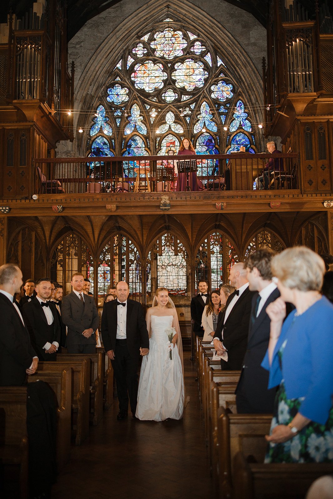 A wedding ceremony inside a historic church with stained glass windows, a wedding couple walking down the aisle, surrounded by formally dressed guests.