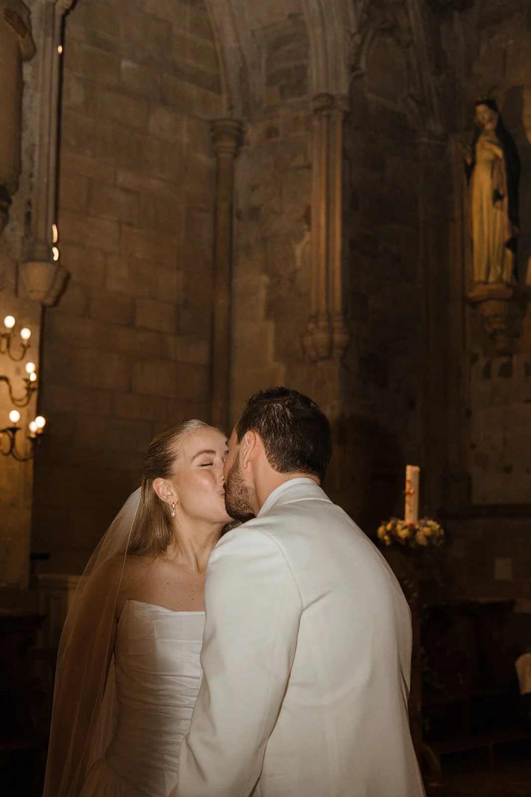 A bride and groom sharing a kiss inside a church with stone walls, candles, and religious statues.