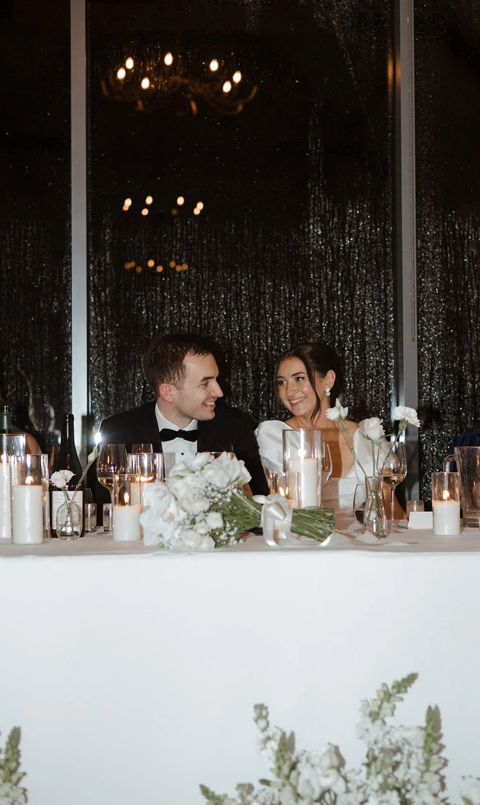 A smiling bride and groom seated at a wedding reception table with candles and floral arrangements, in front of a dark, glittering curtain backdrop.