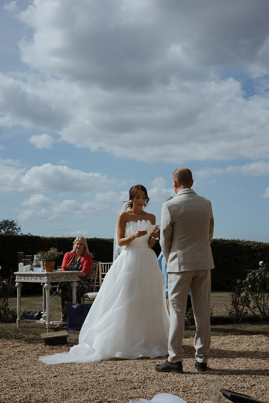 A bride and groom exchanging vows outdoors under a cloudy sky, with a woman sitting at a table nearby.