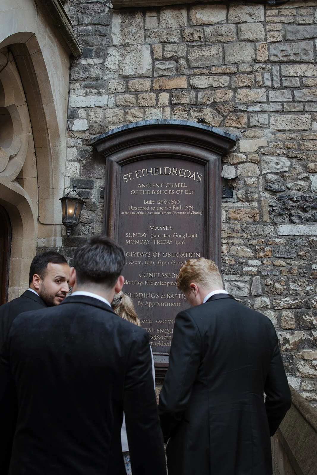 A large wooden informational sign mounted on a stone wall outside St. Etheldreda's church, with several men in suits standing in front of it, engaged in conversation.