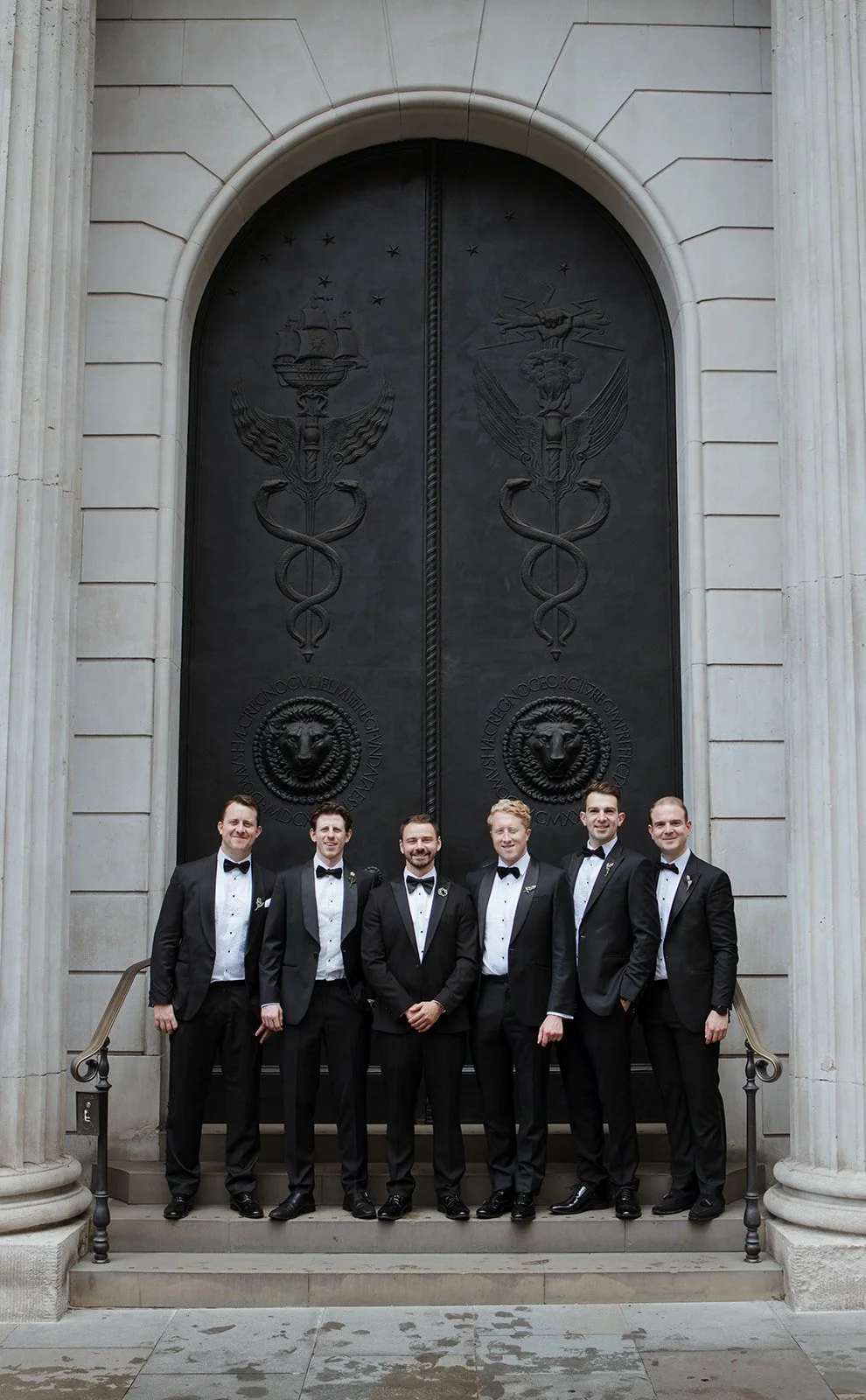 Six men in tuxedos standing in front of a large ornate door with emblems and Latin inscriptions, on the steps of a grand building.