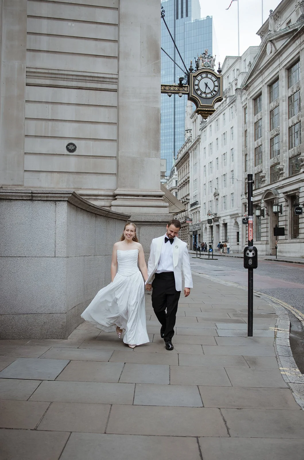 A couple in wedding attire walking hand in hand on a city sidewalk, with tall historic and modern buildings in the background and a clock hanging from a building above them.