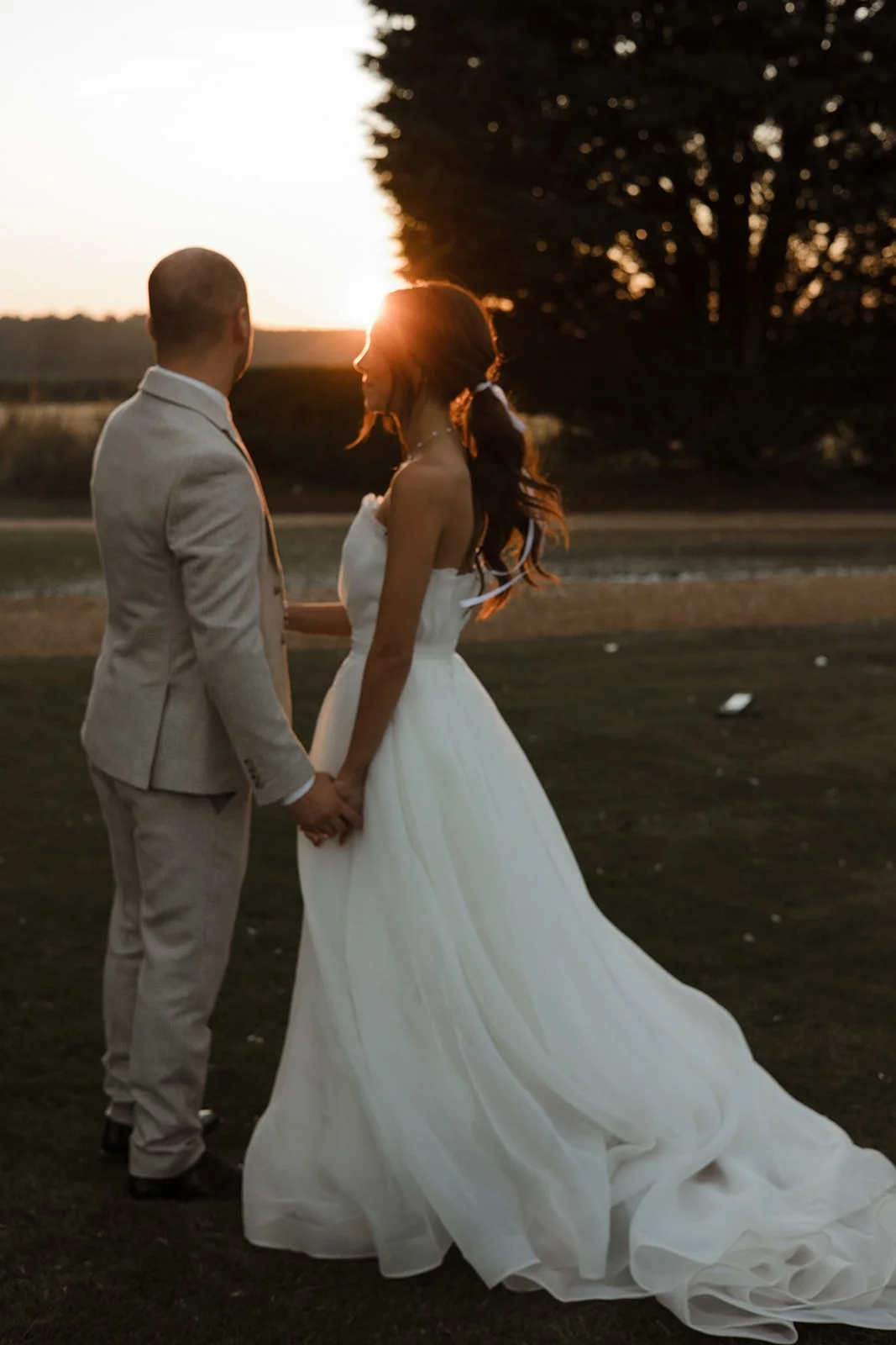 A bride and groom holding hands outdoors at sunset, with the sun setting behind a tree, creating a romantic atmosphere.