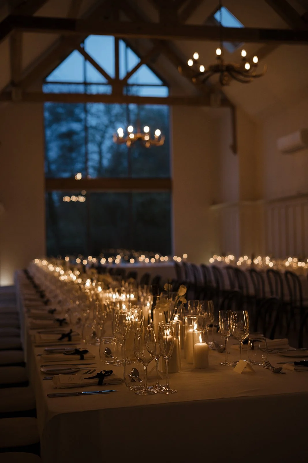 A long banquet table set for a formal dinner in a dimly lit room with candles and wine glasses, with large windows and chandeliers overhead.