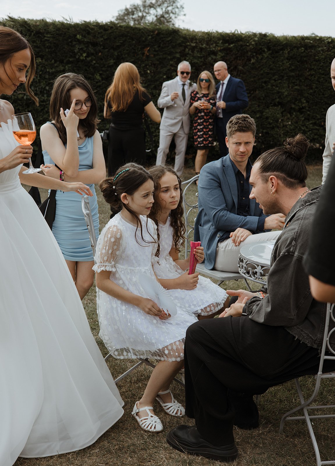 Group of people at an outdoor event, with two young girls in white dresses sitting on chairs, adults standing nearby, and others in the background, some holding drinks.