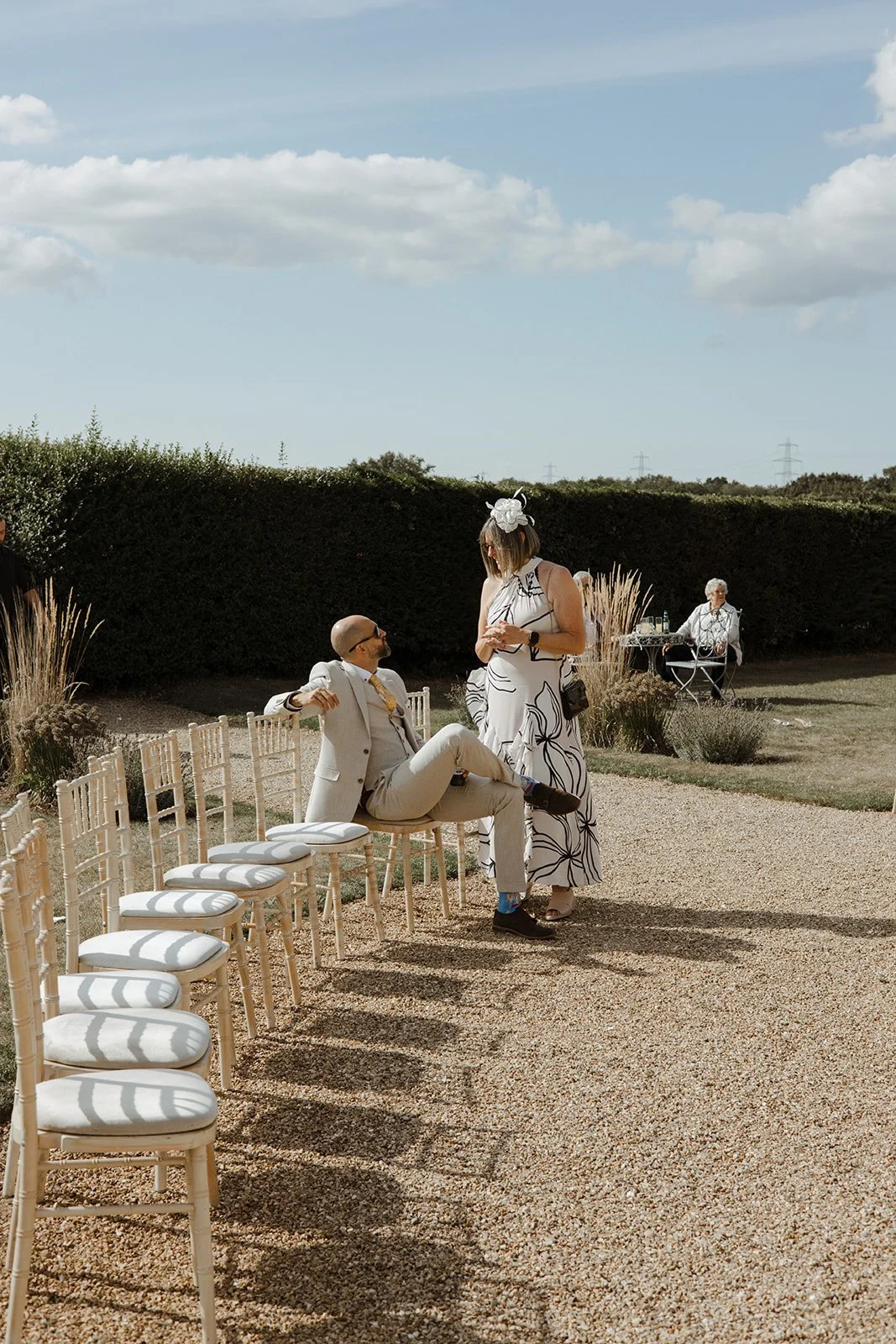 A man in a light-colored suit and sunglasses sitting on a white chair, talking to a woman in a white dress with black floral patterns and a large white hat, at an outdoor daytime event with clear skies and chairs arranged on a gravel path.