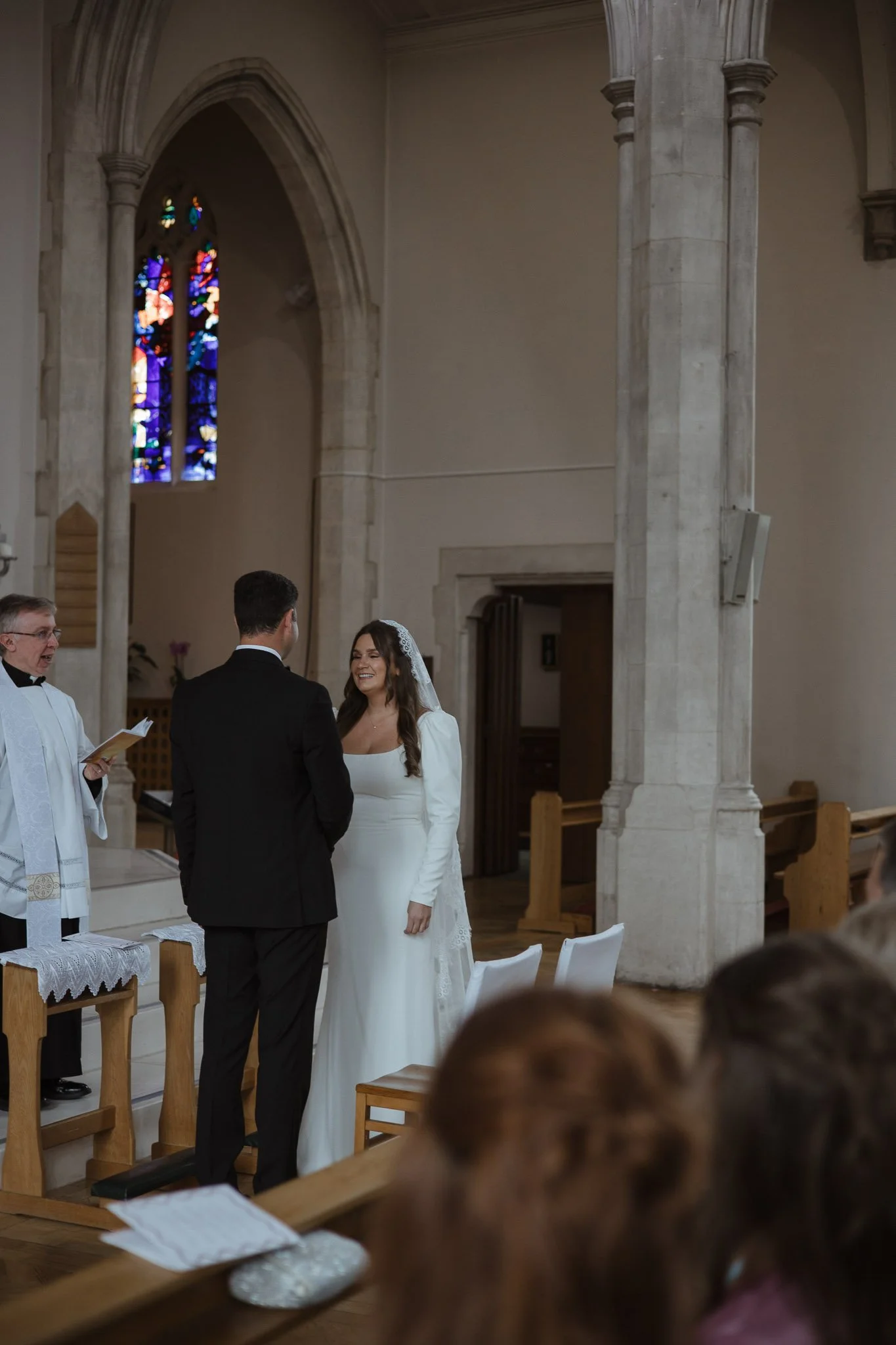 A wedding ceremony taking place inside a church with stained glass windows. The bride and groom are standing facing each other, with the officiant reading from a book. The bride is in a white dress and veil, smiling at the groom, who is in a black su