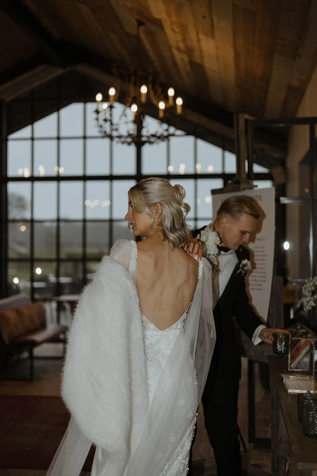 A bride with blonde hair in loose curls, wearing a white wedding gown with a low back and delicate lace details, stands inside a rustic venue with large windows and a chandelier. A groom in a black tuxedo with a bow tie stands nearby, looking at item