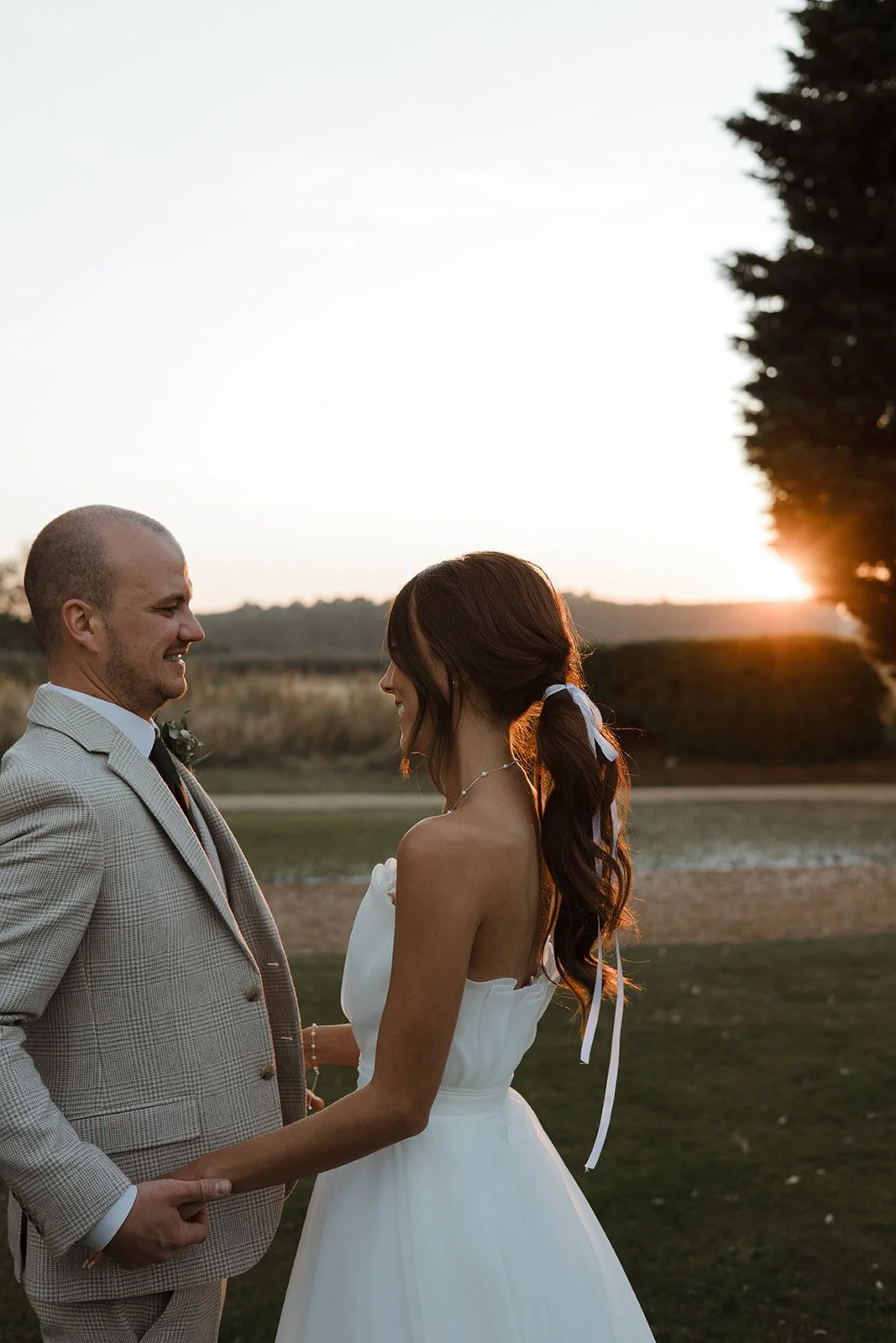 A bride and groom holding hands during sunset outdoors, smiling at each other.