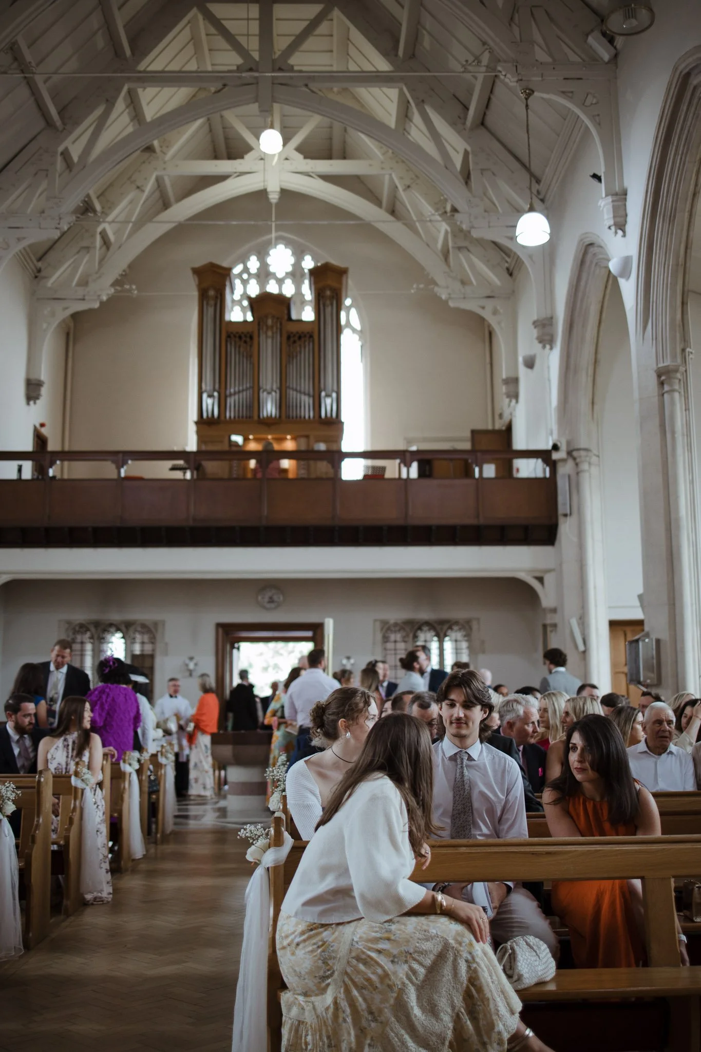 People attending a wedding ceremony inside a church with high vaulted ceiling, wooden pews, and an organ at the front. Some guests are seated, while others are standing and socializing.
