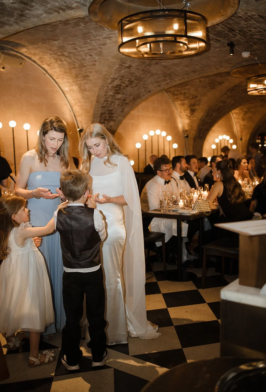 A group of well-dressed people dining in a dimly lit, elegant restaurant with arched brick ceilings and modern circular chandeliers, enjoying a social event.