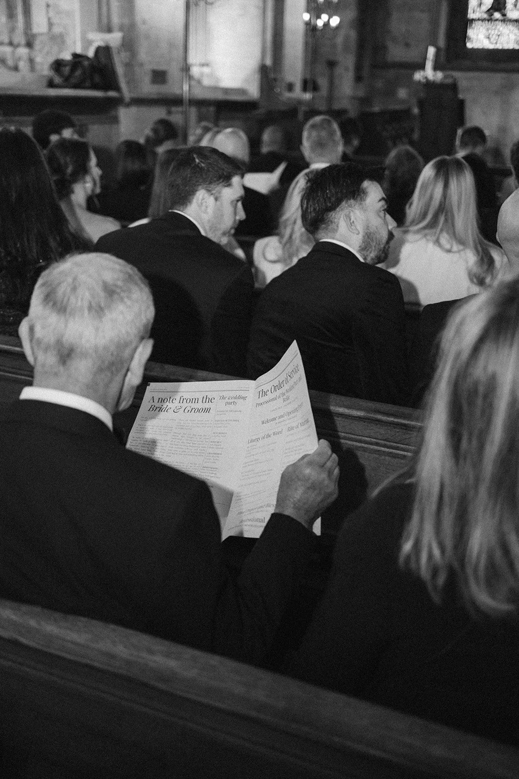 People attending a wedding service in a church, some sitting and reading programs, dressed in formal attire.