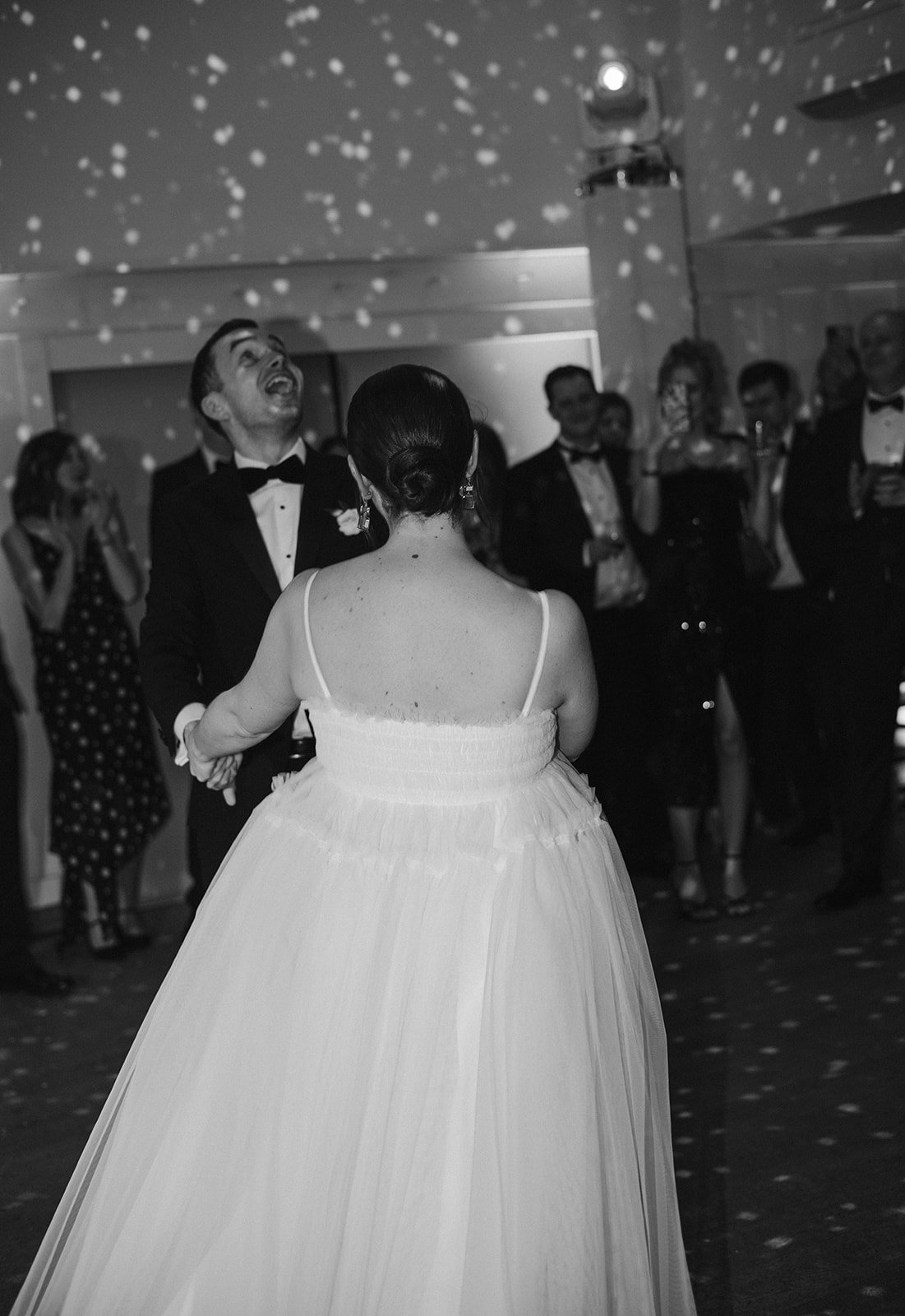 A black and white photo of a bride and groom dancing at their wedding reception with guests watching, decorated with hanging lights.