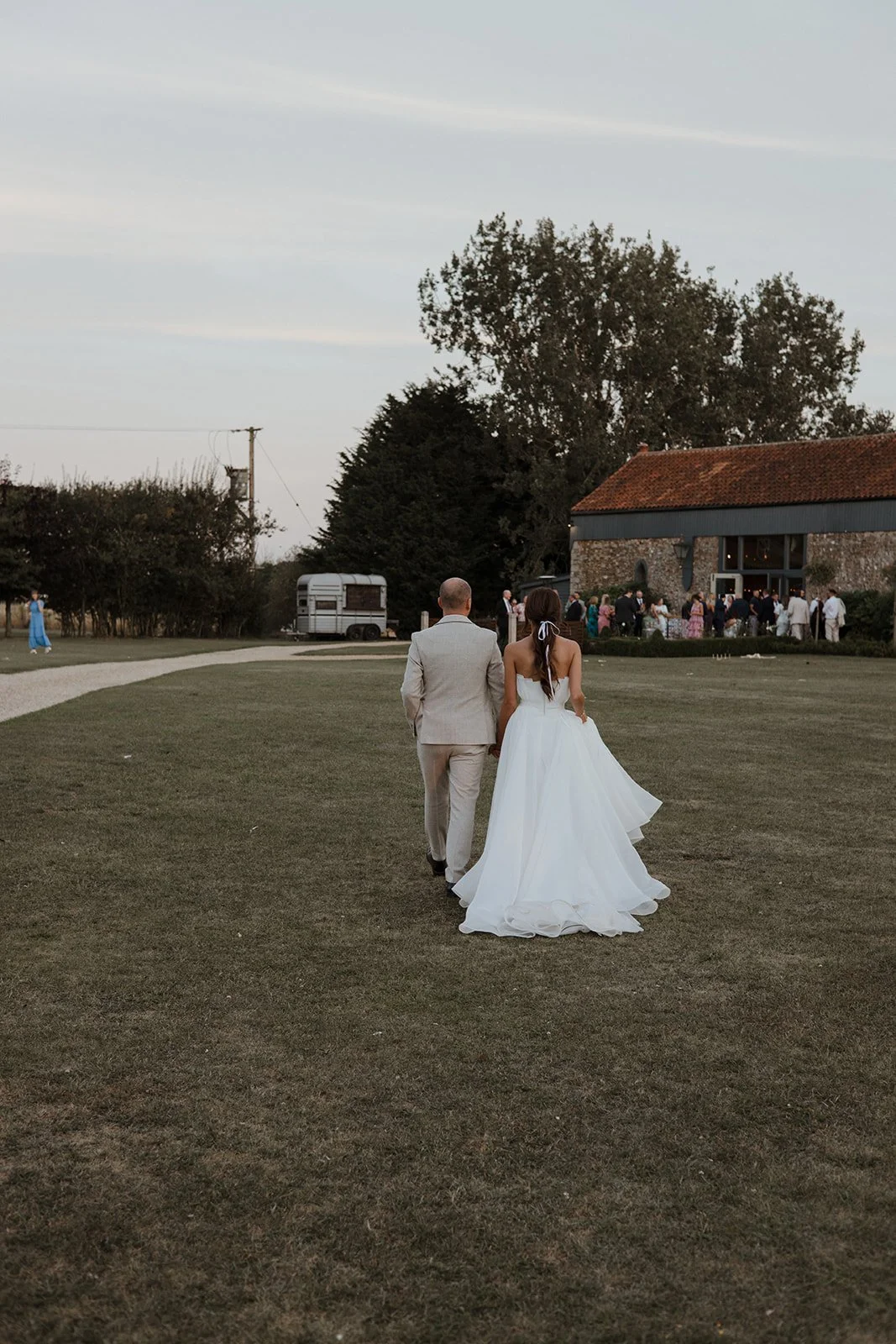 A bride and groom walking on a grassy field during a wedding reception at dusk, with guests mingling near a rustic barn in the background.