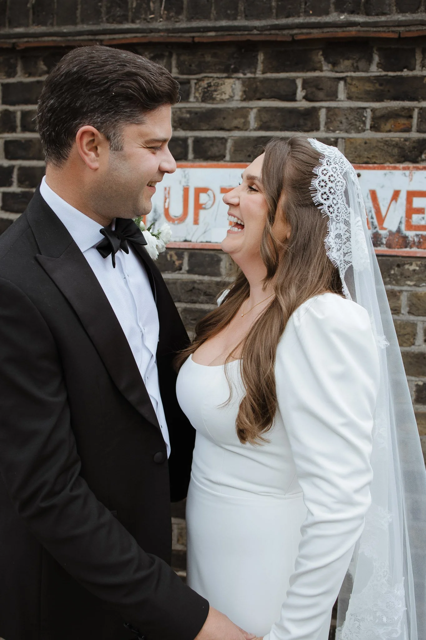 A wedding couple smiling and looking at each other, with a brick wall and vintage sign in the background.