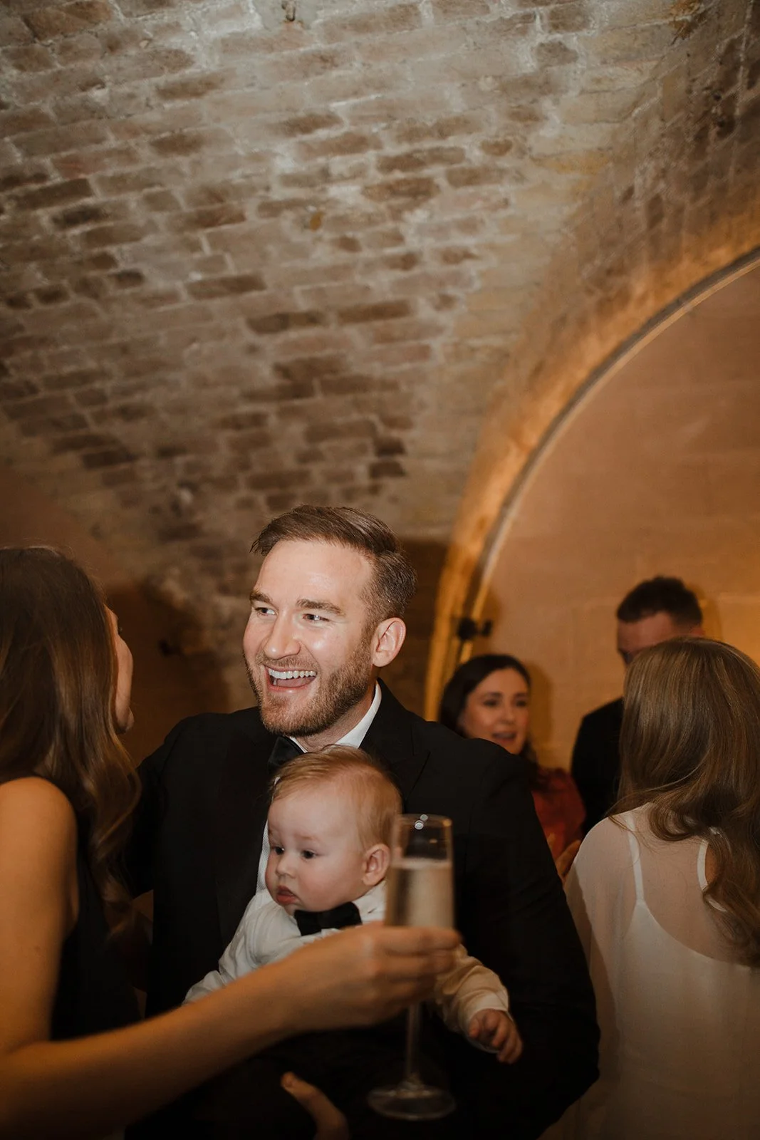 Man in tuxedo holding baby and talking to woman at a social gathering in a brick-walled room.