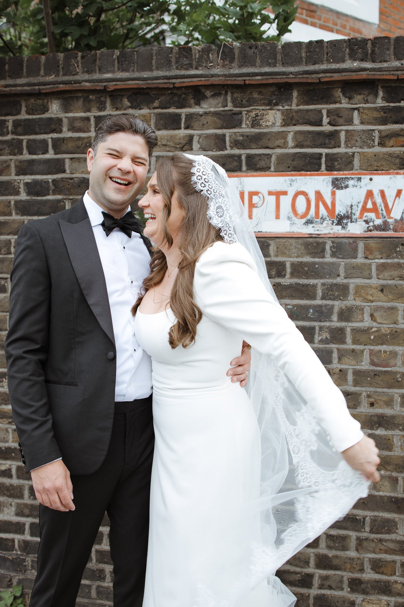 A smiling couple dressed in wedding attire, standing in front of a brick wall with a partially visible street sign.