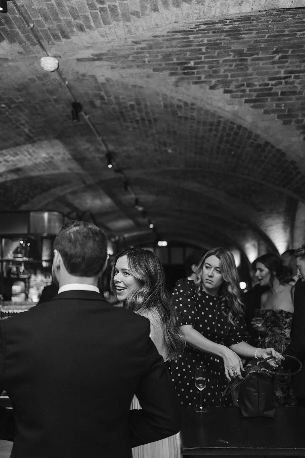 Group of people socializing at a bar or restaurant with arched brick ceiling, some holding drinks, in black and white.