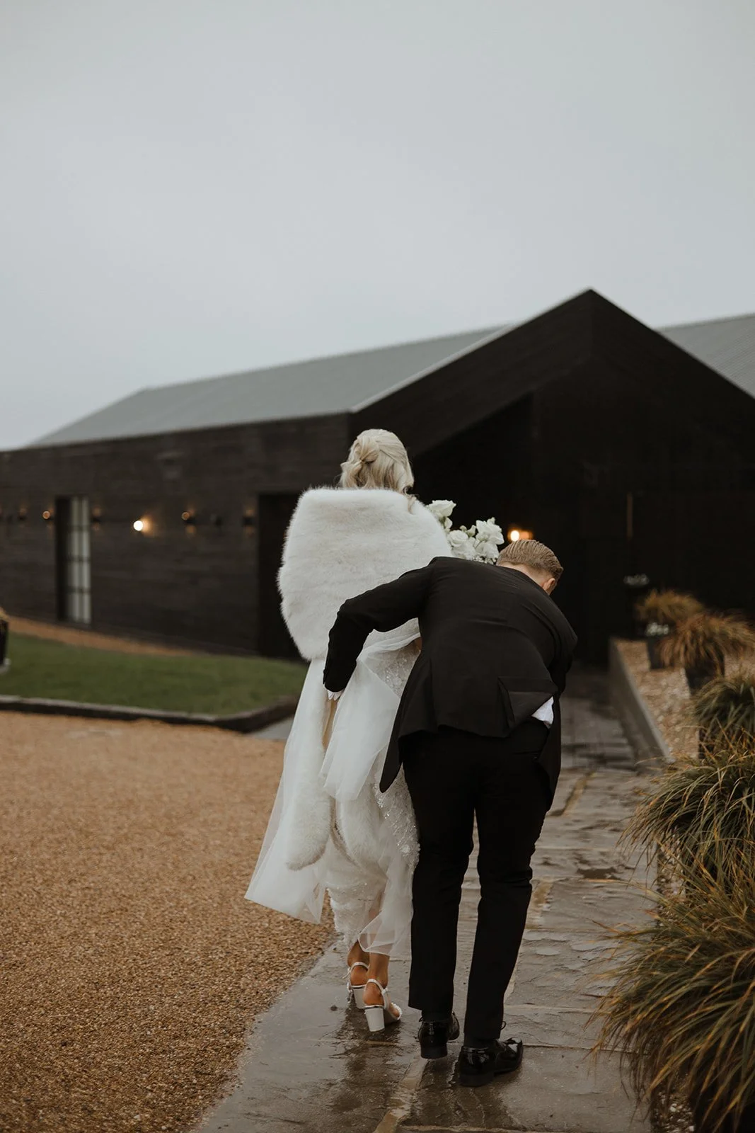Bride in white wedding dress with fur stole walking on wet path, accompanied by a man in dark suit, outside near a dark wooden building under overcast sky.
